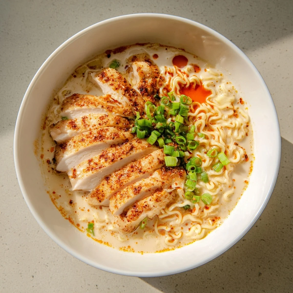 Steaming bowl of fiery chicken ramen, garnished with fresh green onions; a comforting, spicy meal.