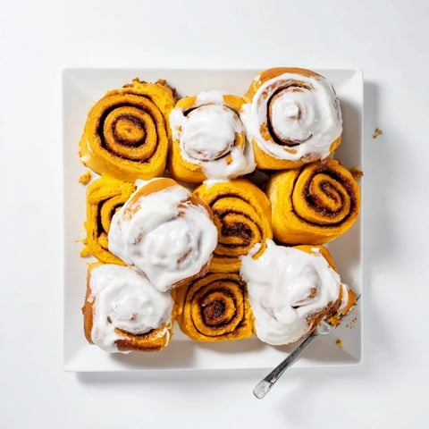 Golden-brown Pumpkin Cinnamon Rolls are drizzled with cream cheese frosting on a rustic wooden board, ready for breakfast or dessert.  