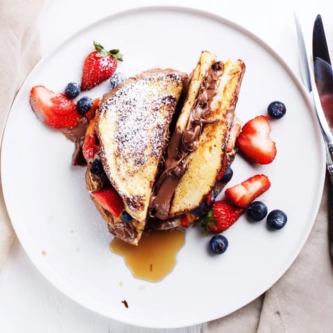 Warm golden Nutella Brioche French toast dusted with powdered sugar, fresh berries, and a drizzle of maple syrup on a white plate.